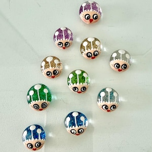 May include: A collection of eleven round, decorative glass cabochons with a whimsical face design. Each cabochon features a different color scheme, including purple, gold, green, and blue, with glitter accents and a red lip.