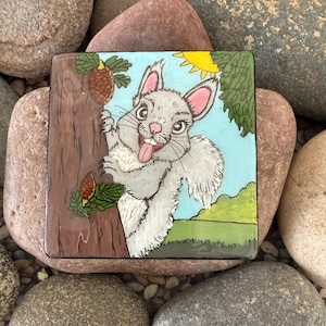 Puede incluir: Azulejo de cerámica cuadrado con una ardilla de dibujos animados en un árbol. La ardilla es gris con orejas y lengua rosas. El fondo es azul claro con un sol amarillo y follaje verde. El azulejo tiene un borde marrón.