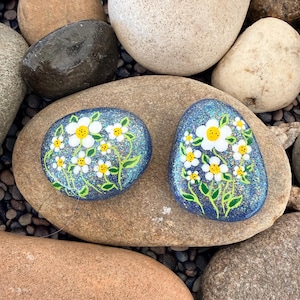 May include: Two hand-painted decorative rocks with a blue glitter background and white daisy floral design. The daisies have yellow centers and green stems and leaves. The rocks are on a larger rock.