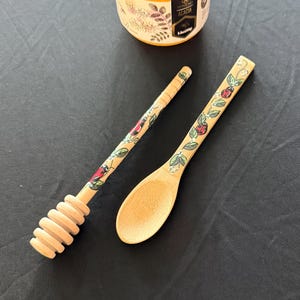 May include: A wooden honey dipper and spoon set with a jar of honey. The dipper has a spiral design, and both utensils feature a painted floral and ladybug pattern. The honey jar has a yellow lid and a label with text.