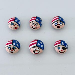 Könnte beinhalten: Sechs runde, handbemalte dekorative Knöpfe mit Cartoon-Gesichtern. Jeder Knopf zeigt ein Design der amerikanischen Flagge auf der Oberseite, mit roten und weißen Streifen und Blau mit weißen Sternen. Die Gesichter haben unterschiedliche Ausdrücke, einschließlich Zwinkern, Lächeln und Überraschung.