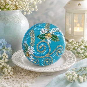 May include: A round, bright turquoise decorative object featuring intricate dot-painted patterns in gold, white, and green. The design includes swirling lines, floral motifs, and small metallic gold beads embedded within the painted surface.