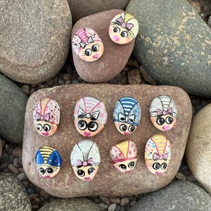 May include: A collection of painted rocks featuring whimsical faces with bows and decorative head coverings. The rocks are painted with various colors, including pink, blue, yellow, and white, and are arranged on larger stones.