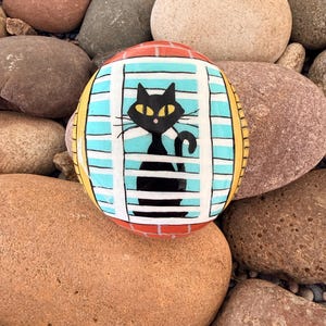 May include: A hand-painted rock featuring a black cat looking out a window. The window has a blue background with white stripes, and the top is painted to look like brick. The rock is surrounded by other rocks.