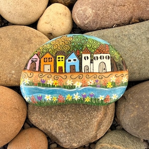 May include: A hand-painted rock featuring a colorful village scene. The artwork depicts a row of houses with varying colors and architectural details, set against a backdrop of a blue river and green foliage. The rock is surrounded by other natural stones.