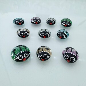 May include: A collection of hand-painted, decorative rocks, each featuring a black base with a stylized ladybug design. The ladybugs have white eyes, a red heart-shaped mouth, and colorful wings in shades of green, blue, gray, and purple.