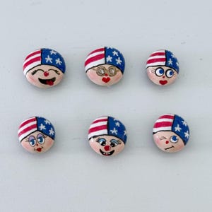 May include: Six hand-painted, round decorative stones. Each stone features a face with a patriotic design: a blue cap with white stars and red and white stripes. The faces have different expressions, including winks, smiles, and glasses.