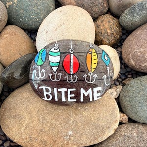 May include: A painted rock with a fishing theme. The rock features colorful fishing lures and the words "BITE ME" in white lettering. The rock is surrounded by other rocks of various sizes and colors.