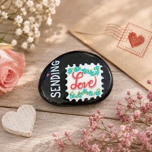 May include: A smooth, black, oval-shaped stone painted with the white text "SENDING" and a white, postage stamp-shaped graphic featuring blue flowers and the red script word "Love" in the center, with a small red heart above the text.
