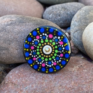 May include: A round, black decorative object with a colorful mandala design. The mandala features blue, pink, green, white, and yellow dots arranged in concentric circles. A small, gold-colored embellishment is in the center.