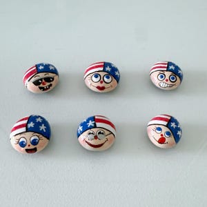 May include: Six painted rocks with faces, each featuring a red and white striped design and blue star accents. The faces have various expressions, including sunglasses, smiles, and winks. The rocks are arranged on a white surface.