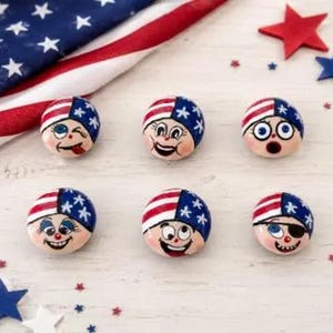Pode incluir: Seis botões redondos de madeira com rostos pintados à mão e expressões variadas. Cada botão retrata uma personagem usando um chapéu com o padrão da bandeira americana, composto por listras vermelhas e brancas e um campo azul com estrelas.