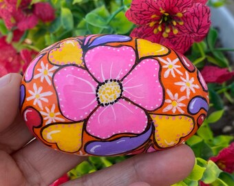 Puede incluir: Piedra pintada a mano, de forma ovalada, con una gran flor rosa con centro amarillo y puntos blancos. La piedra tiene un fondo naranja con diseños en forma de corazón amarillos, morados y rojos y pequeñas flores blancas en forma de margarita. Un artículo decorativo único.