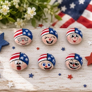 Pode incluir: Seis pedras redondas pintadas à mão com rostos usando chapéus com o design da bandeira americana. Cada pedra exibe uma expressão facial única, incluindo óculos, olhos grandes e sorrisos, nas cores vermelho, branco e azul com detalhes de estrelas.