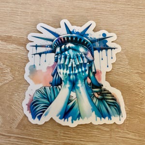 May include: A sticker featuring a watercolor illustration of the Statue of Liberty with her hands covering her face, as if crying. The statue is depicted in shades of blue and green, with a pink and orange background.