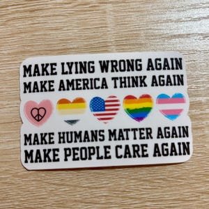 May include: White sticker with black text that reads "Make Lying Wrong Again", "Make America Think Again", "Make Humans Matter Again", and "Make People Care Again". The sticker features five hearts with different symbols: a peace sign, a transgender flag, an American flag, a rainbow flag, and a gender-neutral flag.
