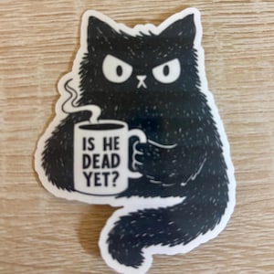 May include: A black cat sticker with a grumpy expression, holding a coffee mug. The mug has the text "IS HE DEAD YET?" in black letters. The sticker is on a light brown wooden surface.