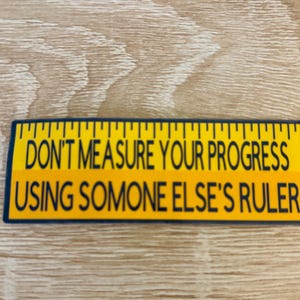 May include: A yellow and black ruler-shaped sticker with the text "DON'T MEASURE YOUR PROGRESS USING SOMEONE ELSE'S RULER". The sticker has a blue border and is on a light brown wooden surface.