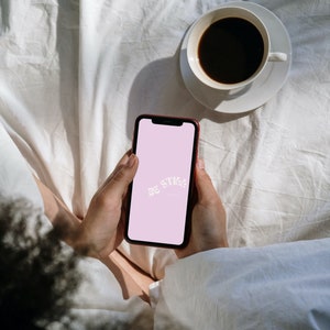 Op de afbeelding: Een rode smartphone met de tekst "Be Still" op een roze scherm wordt vastgehouden. Een witte koffiekop met donkere koffie staat op een schotel. De sc&egrave;ne speelt zich af op een wit, gekreukt laken.
