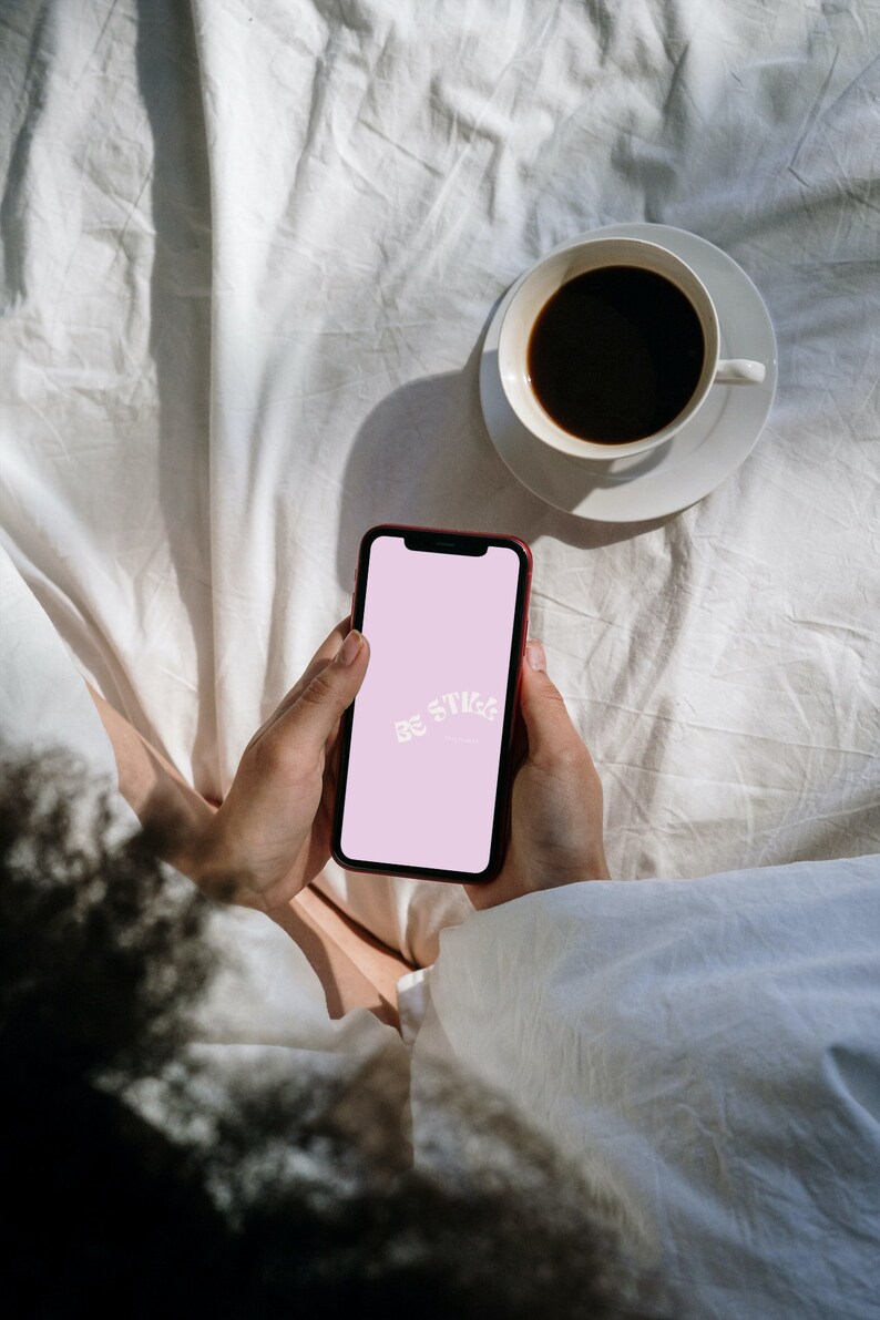 Op de afbeelding: Een rode smartphone met de tekst "Be Still" op een roze scherm wordt vastgehouden. Een witte koffiekop met donkere koffie staat op een schotel. De sc&egrave;ne speelt zich af op een wit, gekreukt laken.