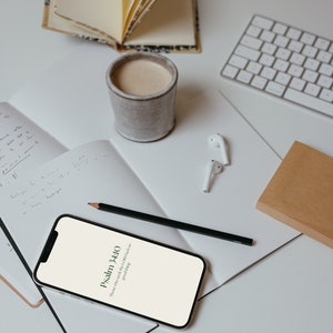 Op de afbeelding: Een bovenaanzicht van een werkruimte met een smartphone met tekst uit Psalm 34:10, een kop koffie, een notitieboek, een potlood, een toetsenbord en een klein open boek. De items zijn gerangschikt op een wit oppervlak.
