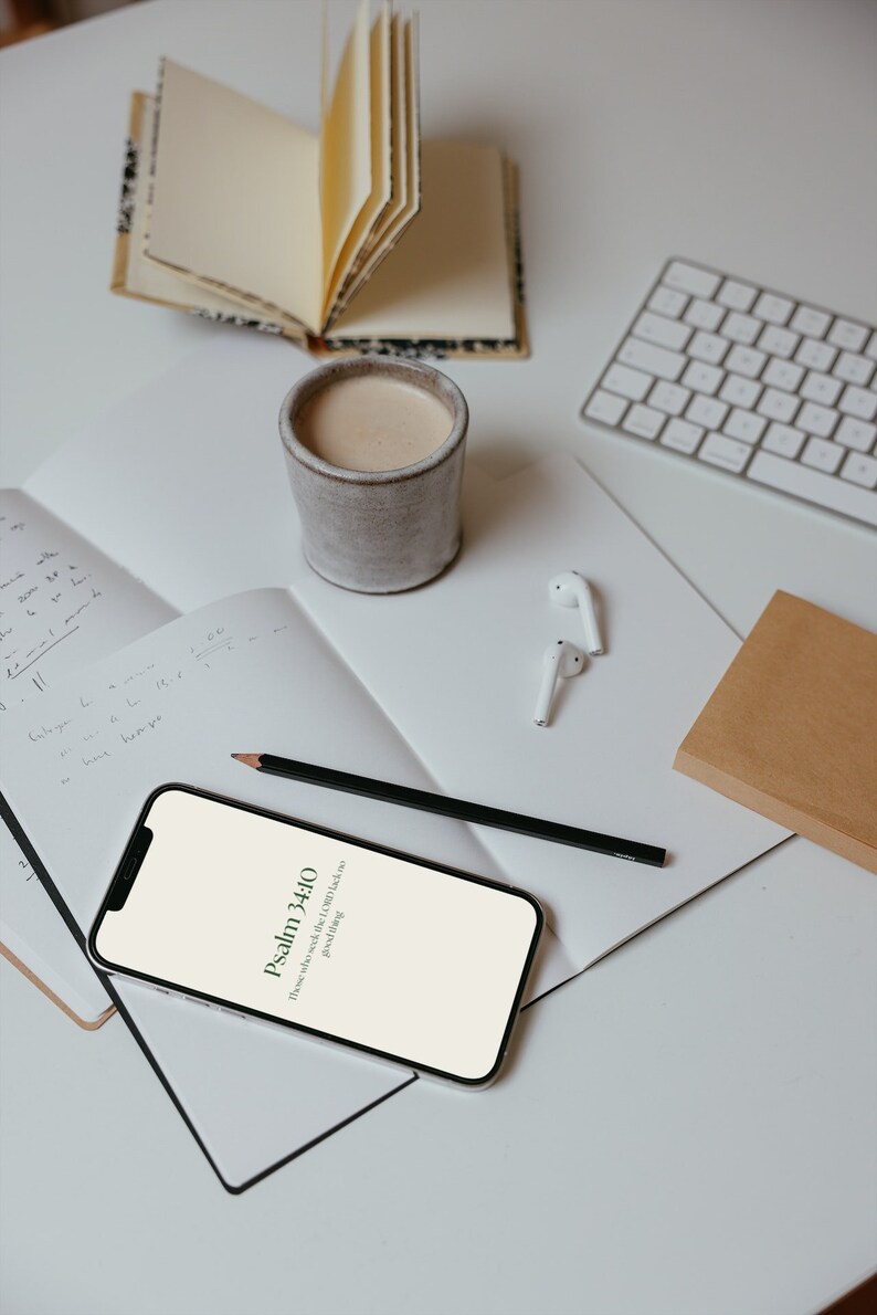 Op de afbeelding: Een bovenaanzicht van een werkruimte met een smartphone met tekst uit Psalm 34:10, een kop koffie, een notitieboek, een potlood, een toetsenbord en een klein open boek. De items zijn gerangschikt op een wit oppervlak.