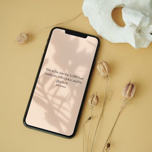 Op de afbeelding: Een zwarte smartphone toont een lichtroze scherm met de tekst "This is the day the LORD has made; we will rejoice and be glad in it. PSALM 118:24." Gedroogde bloemen en een witte steen zijn op een gele achtergrond geplaatst.