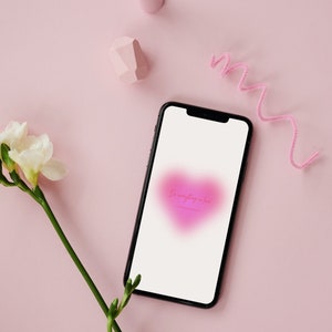 Op de afbeelding: Een zwarte smartphone toont een roze hart met de tekst "Do everything in love" op een roze achtergrond. De afbeelding bevat een witte bloem, roze geometrische vormen en een roze pijpenrager.