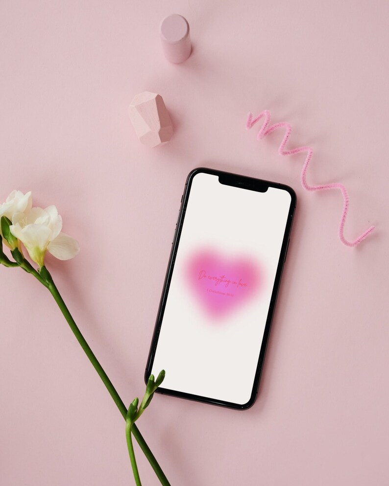 Op de afbeelding: Een zwarte smartphone toont een roze hart met de tekst "Do everything in love" op een roze achtergrond. De afbeelding bevat een witte bloem, roze geometrische vormen en een roze pijpenrager.