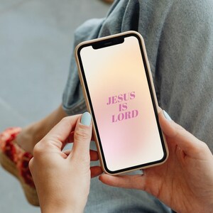 Op de afbeelding: Een smartphone met een beige hoesje toont een gradi&euml;nt achtergrond met de tekst "JESUS IS LORD" in roze. De telefoon wordt vastgehouden door een persoon die een blauwe spijkerbroek en lichtblauwe nagellak draagt.
