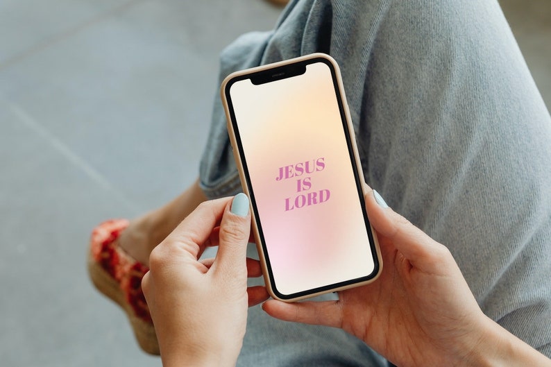 Op de afbeelding: Een smartphone met een beige hoesje toont een gradi&euml;nt achtergrond met de tekst "JESUS IS LORD" in roze. De telefoon wordt vastgehouden door een persoon die een blauwe spijkerbroek en lichtblauwe nagellak draagt.