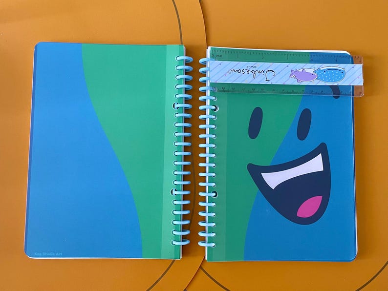 Puede incluir: Una libreta de espiral azul y verde con una cara sonriente en la portada. La libreta tiene una encuadernaci&oacute;n de pl&aacute;stico azul y una regla con el texto "Jungle Scout" en ella.