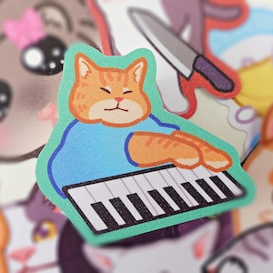 以下が含まれることがあります： オレンジ色の猫がキーボードを弾いているステッカー。猫は青いシャツを着ており、キーボードは白黒の鍵盤です。ステッカーは緑色の背景です。