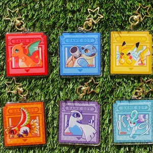 Könnte beinhalten: Sechs bunte Schlüsselanhänger mit Pokémon-Charakteren aus der Game Boy-Videospielserie. Jeder Schlüsselanhänger hat einen durchsichtigen Plastikrahmen mit einem bunten Bild eines Pokémon-Charakters und dem Text "Game Boy".