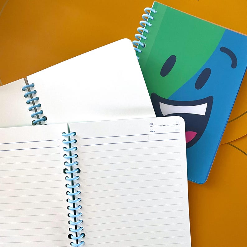 Puede incluir: Dos cuadernos de espiral, uno con una cubierta blanca y el otro con una cubierta verde, azul y rosa con una cara sonriente. El cuaderno con la cara sonriente tiene las palabras "NO." y "Fecha" impresas en la cubierta.