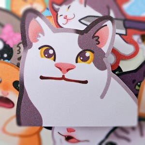 以下が含まれることがあります： 頭の上に灰色の斑点があり、黄色い目をした白い猫。猫は口を少し開けて驚いた表情をしている。猫は光沢のある仕上げのステッカーです。