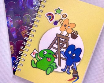 May include: A yellow notebook with a spiral binding features a cartoon illustration of four characters. The characters are a green creature with a tongue sticking out, a blue four with a paintbrush, a yellow four on a ladder, and a purple blueberry. The notebook has a sticker of a rainbow, a star, and a smiley face. The text "KAE ONLINE" is printed on the bottom right corner of the notebook.