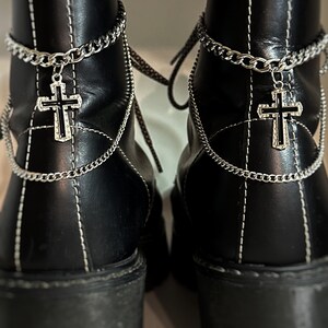 Puede incluir: Un par de botas de cuero negro con costuras blancas y cadenas plateadas con dijes de cruz. Las botas tienen suelas gruesas y un tacón grueso.