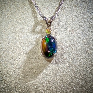May include: A silver chain necklace with a pendant featuring a black opal gemstone with green and gold iridescent flashes. The pendant is accented with a smaller green gemstone.