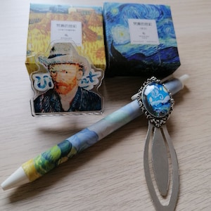 May include: A silver bookmark with a blue and white glass cameo featuring a starry night scene. The bookmark is attached to a pen with a blue and white patterned design. The pen is lying on a wooden surface. The image also includes two small boxes with Japanese text.