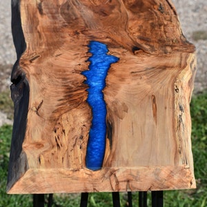 May include: A wooden table top with a live edge and a blue resin river running down the center. The table is supported by four black hairpin legs.