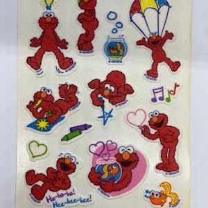 May include: Sheet of vintage Elmo stickers featuring various poses, including Elmo with a lightbulb, sunglasses, a parachute, and a goldfish. Other stickers include musical notes, hearts, and the text "Ha-to-ho! Hee-hee-hee!"