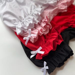 May include: A stack of ruffled bloomers in white, pink, red, and black. Each pair has a ruffled trim and a white satin bow. The bloomers are made of a soft fabric.