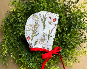 May include: A white baby bonnet with a red ribbon tie. The bonnet has a floral print with small mice and strawberries.