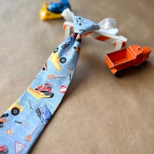 May include: A blue and white tie with a construction theme. The tie features a variety of construction vehicles, including dump trucks, bulldozers, and excavators. The tie is tied around a construction barrier.