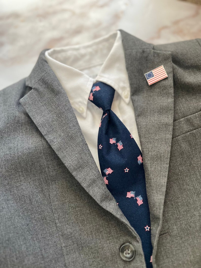 May include: A gray suit jacket with a white collared shirt and a dark blue tie with small American flag designs. A small American flag pin is on the lapel of the jacket.
