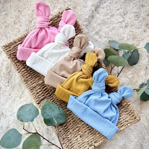 May include: Five different colored baby hats with a knot on top. The hats are in a woven basket and are arranged in a row. The colors of the hats are pink, white, beige, yellow, and blue.