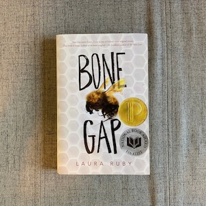 May include: Book cover for "Bone Gap" by Laura Ruby. The cover features a bee on a honeycomb background with the title in black and white lettering. The book is a National Book Award Finalist.
