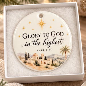 May include: A round white ceramic ornament featuring a gold star, the text "GLORY TO GOD in the highest." and "LUKE 2:14" above a watercolor-style illustration of a desert town with buildings, hills, and a palm tree, presented in a white box.