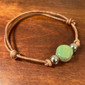 May include: A brown leather bracelet with a green and white circular charm and two silver beads.